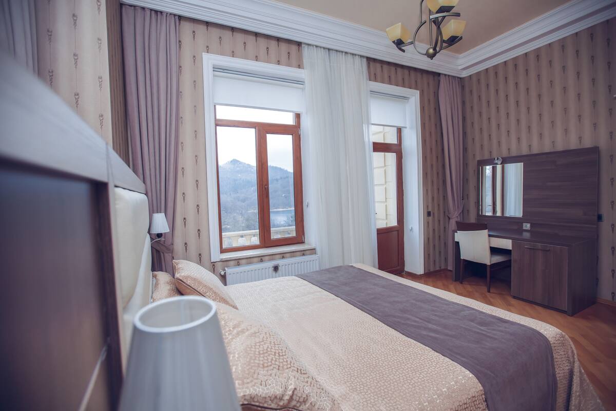 Een slaapkamer met een bed, een bureau, een stoel, een lamp en een groot raam met gordijnen.