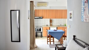 A compact kitchen with wooden cabinets, a dining table with a blue tablecloth, and a bed with white bedding. - Chalkidonos (Heraklion)