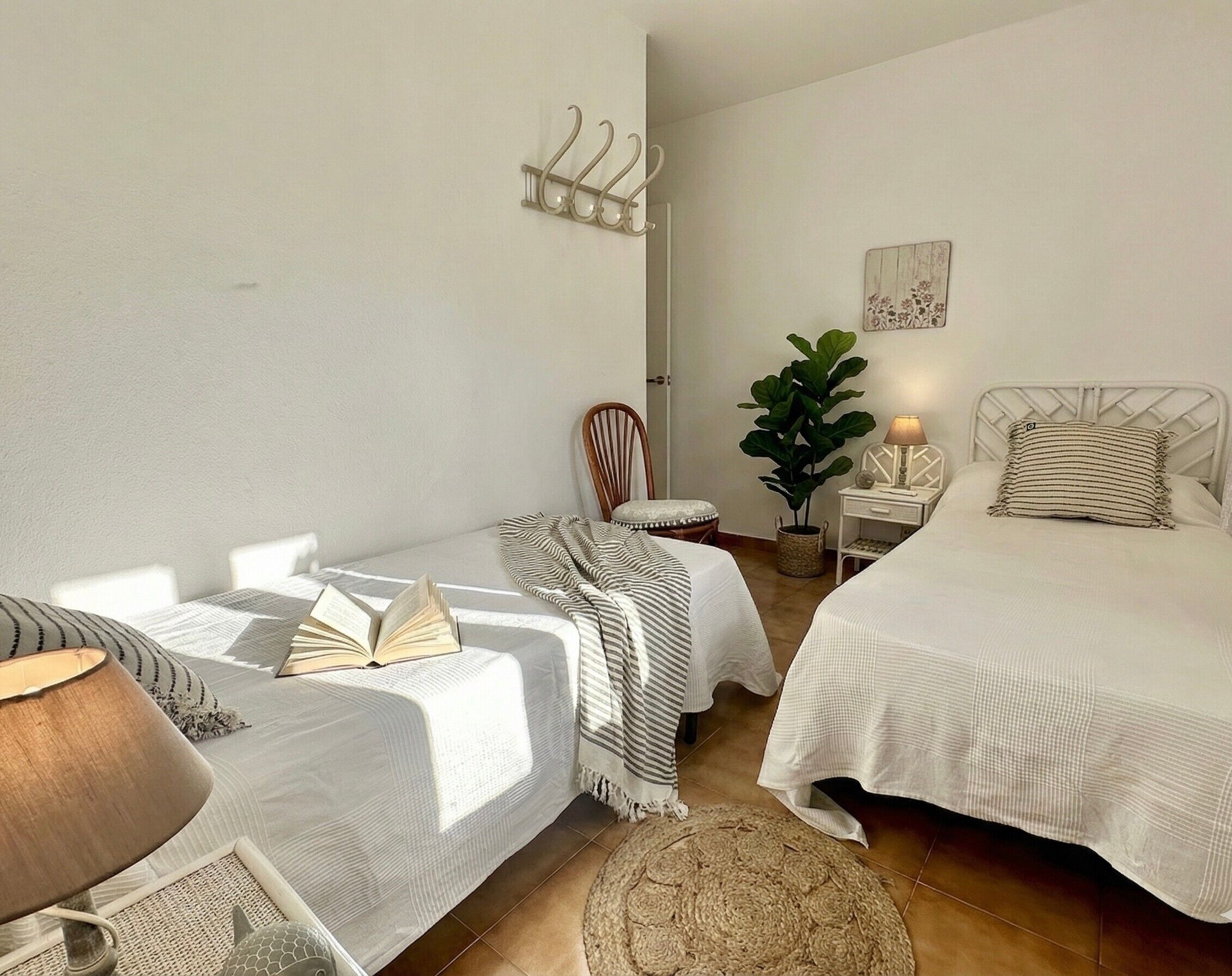 Un dormitorio con dos camas, una silla, una mesita de noche con lámpara, una planta en maceta y un perchero en la pared.