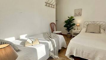 Un dormitorio con dos camas, una silla, una mesita de noche con lámpara, una planta en maceta y un perchero en la pared.