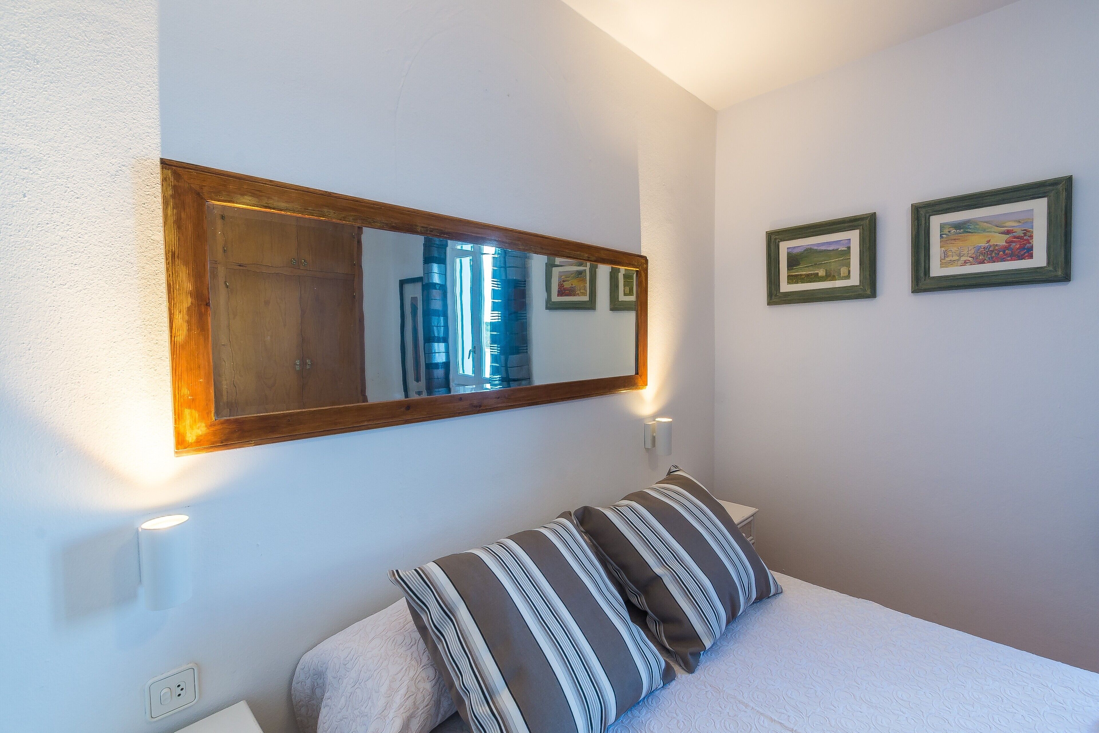 Une chambre avec un grand miroir, un lit agrémenté d’oreillers à rayures et deux tableaux encadrés accrochés au mur.