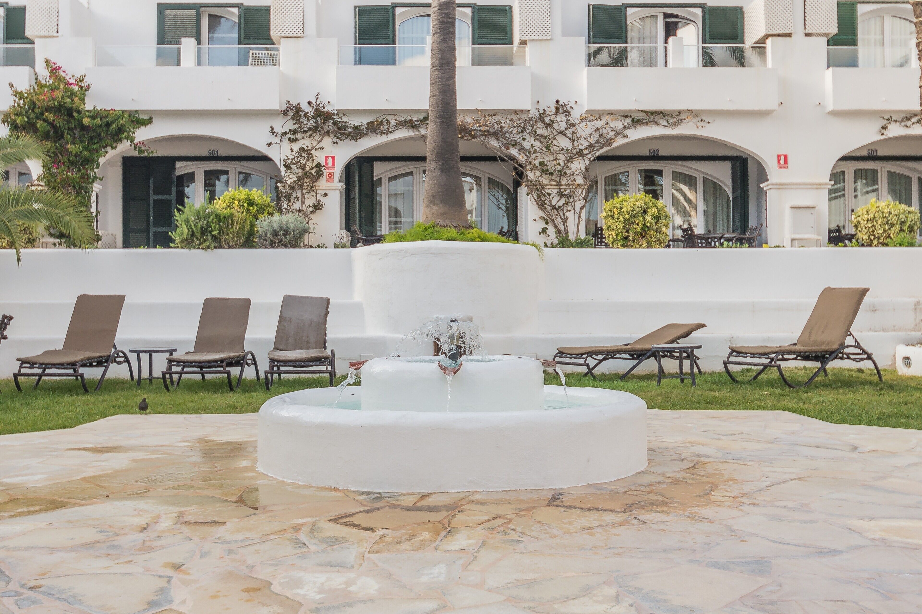 Une cour intérieure avec une fontaine centrale, des chaises longues et un bâtiment blanc doté de balcons.
