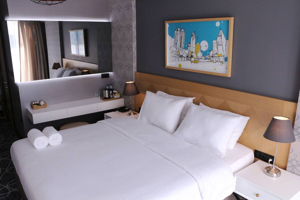 Una habitación de hotel con una cama grande, una mesita de noche con lámpara, un tocador y un cuadro en la pared.