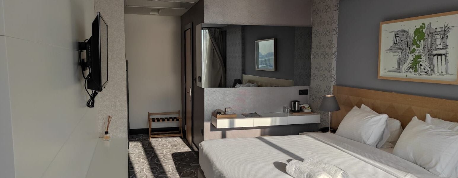 A modern hotel room with a large bed, a bedside table with a lamp, a mirror, and a wall-mounted TV.