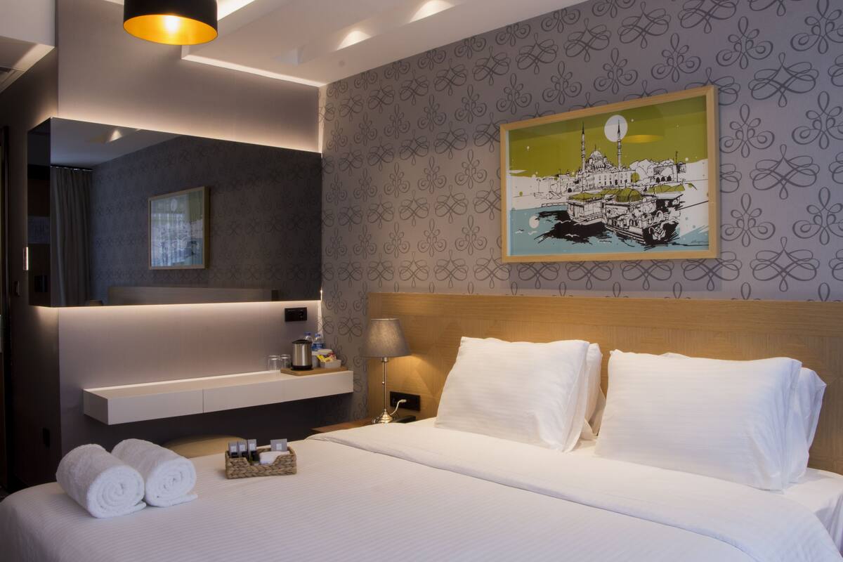 Una habitación de hotel moderna con una cama bien hecha, un cabecero, un estante en la pared con artículos de aseo y una obra de arte enmarcada que representa un paisaje urbano.