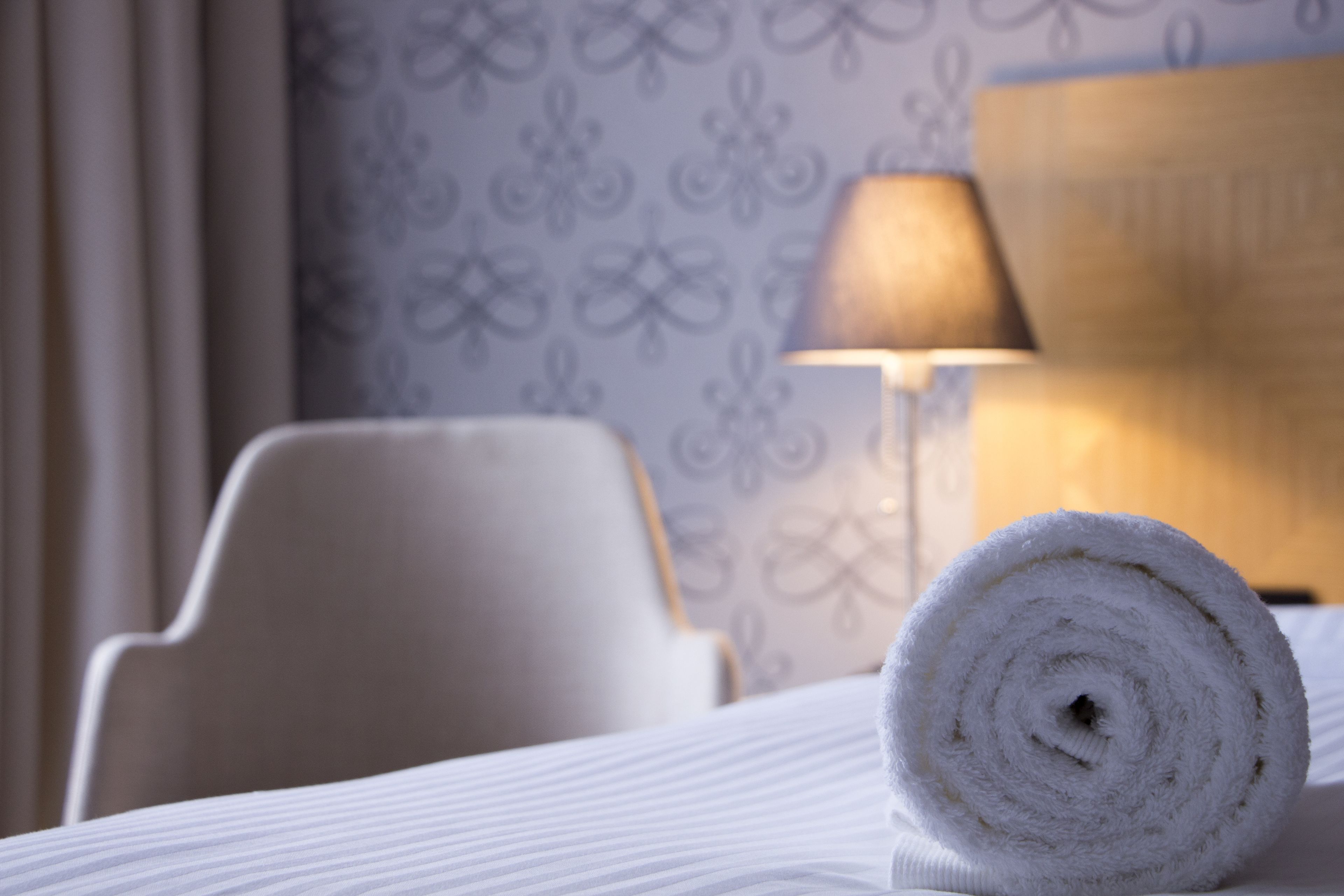 A neatly made bed with a rolled white towel, a white chair, and a lamp in a room with patterned wallpaper.