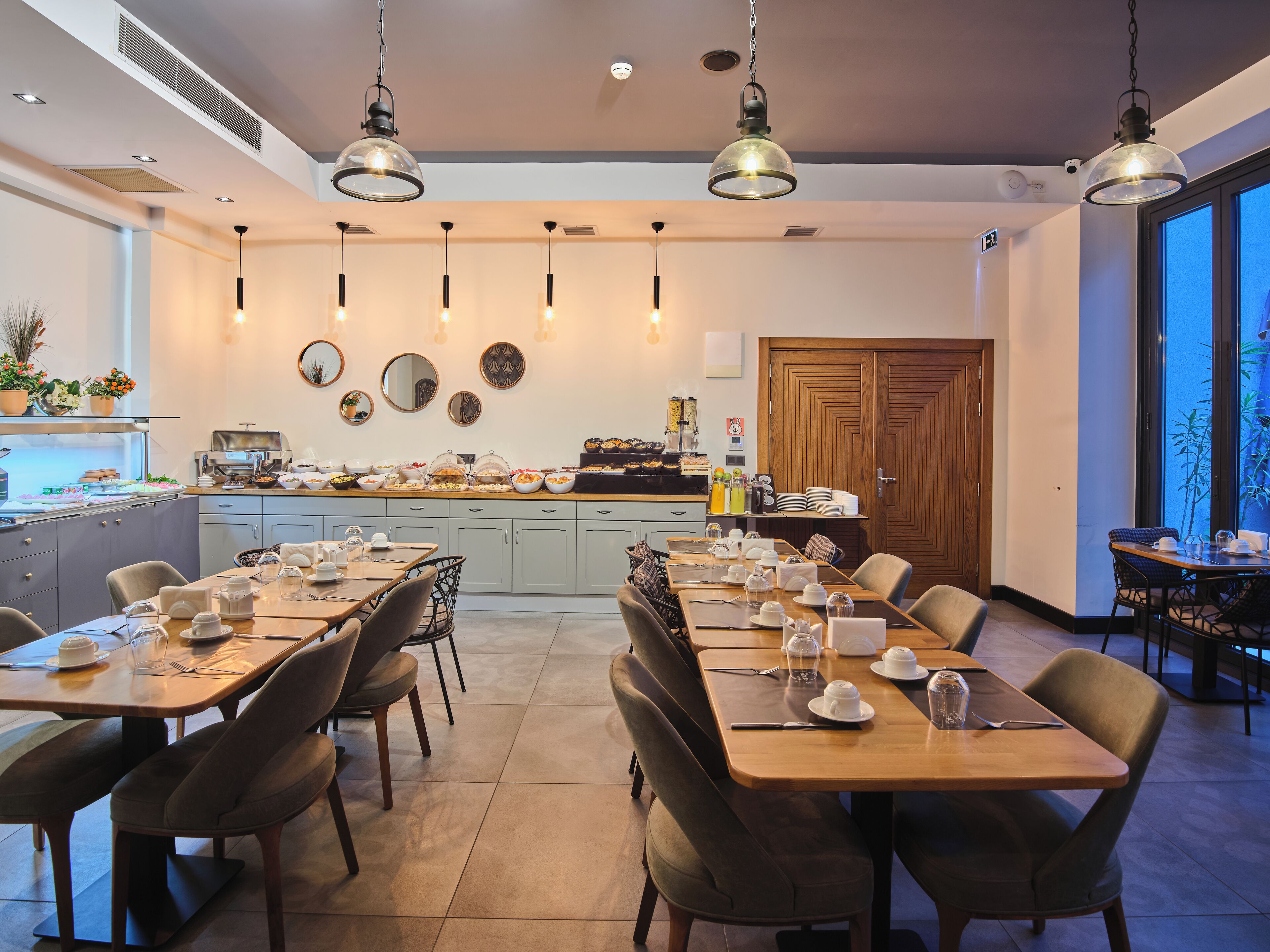 A dining area with multiple tables set for breakfast, a kitchen in the background, and a view of the outdoors through large windows.
