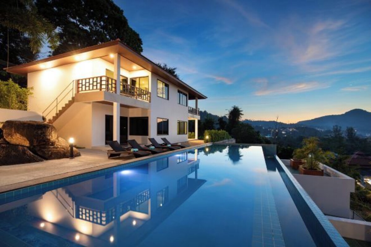 A modern two-story house with a swimming pool, a balcony, and a view of the mountains.