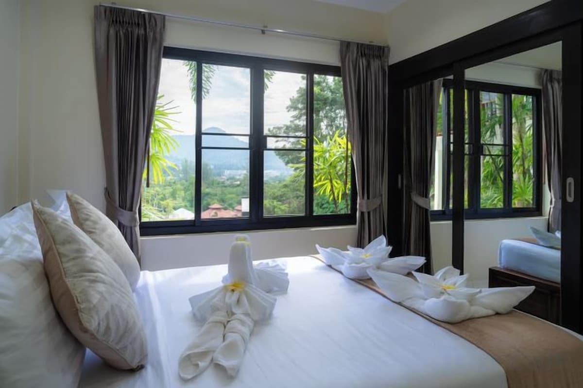 A hotel room with a bed, a window with curtains, and a view of greenery outside.