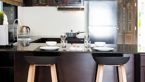 A modern kitchen with dark wooden cabinets, a stainless steel refrigerator, and a central island with stools. - Baan Chom View Kamala (Kamala)