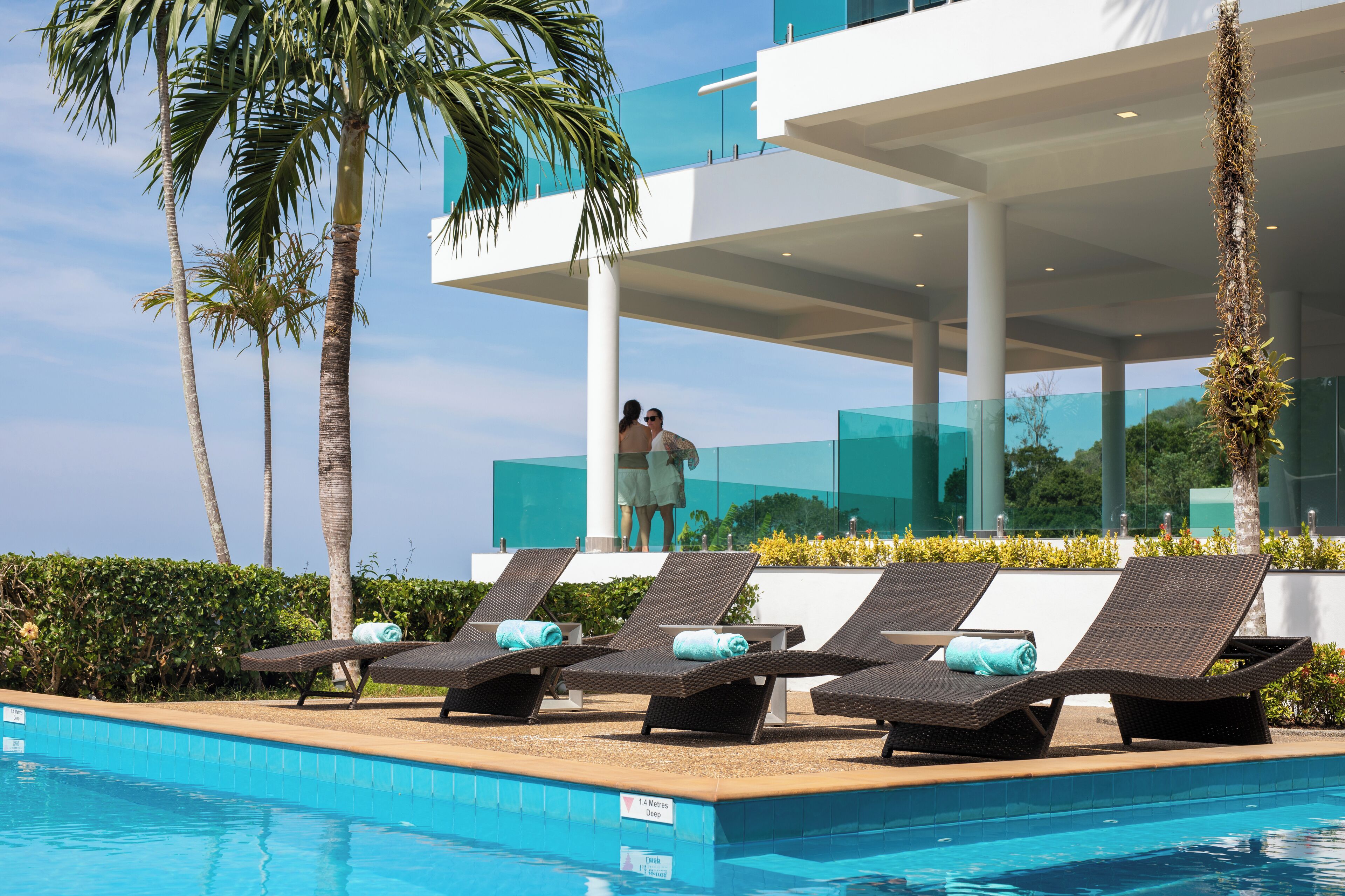 Un área junto a la piscina con sillones de descanso, una piscina azul cristalina y un edificio moderno con balcones de vidrio.