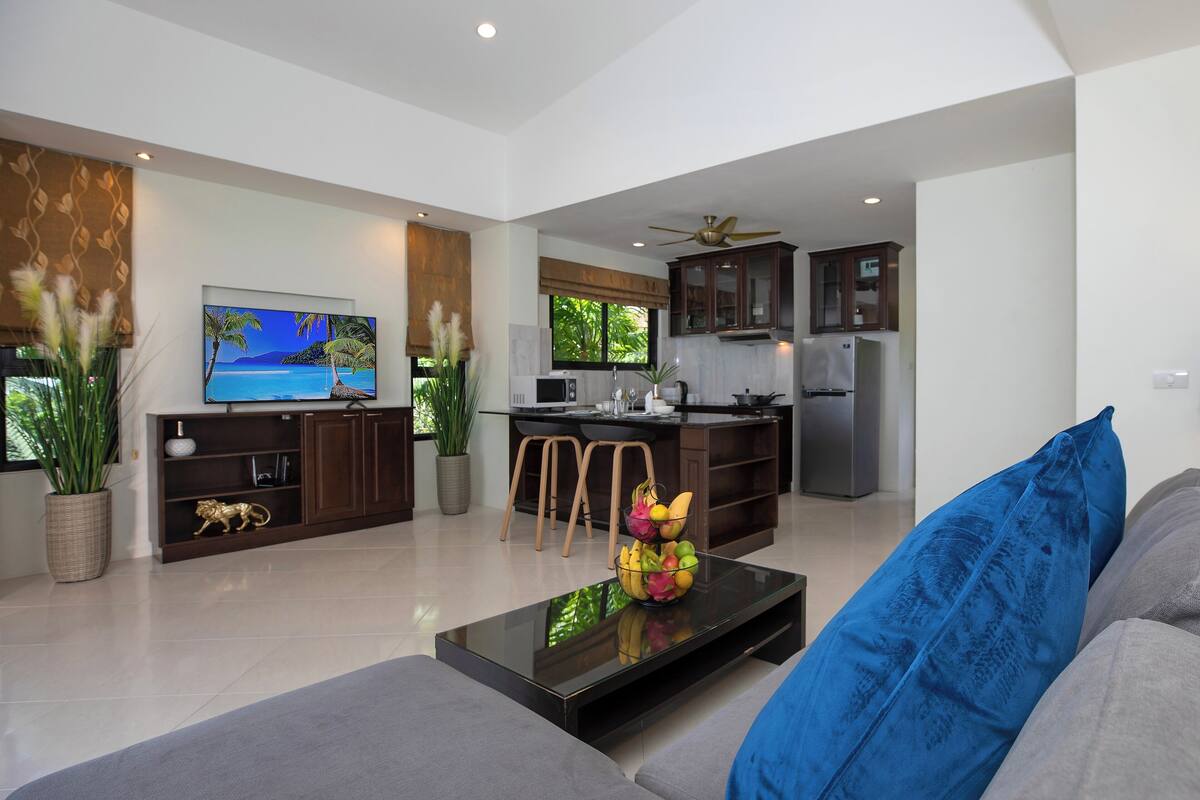 A modern living room with a flat-screen TV, a coffee table with fruit, a sofa with a blue pillow, and a kitchen area with a refrigerator and microwave.