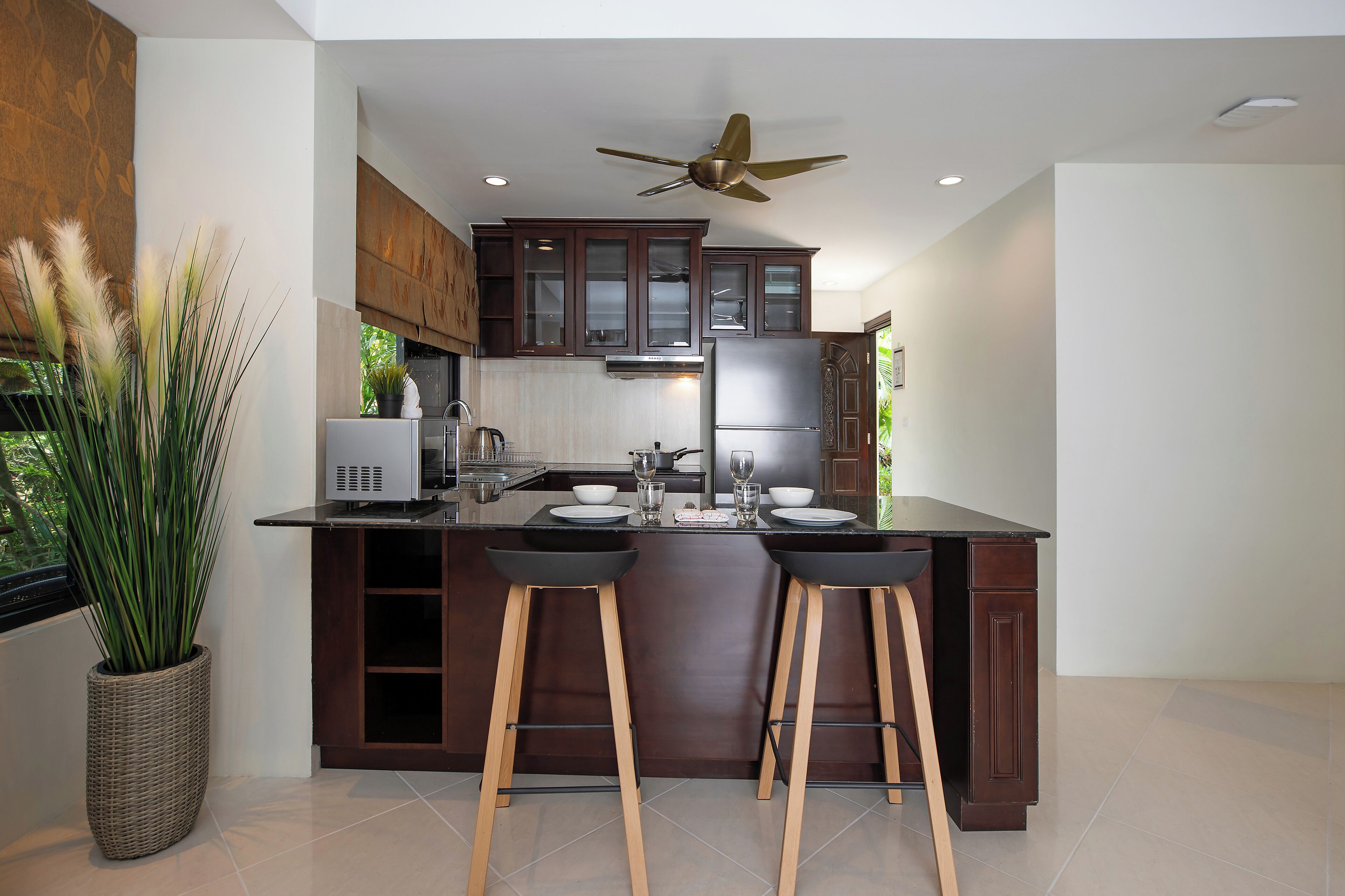 A modern kitchen with dark wooden cabinets, a central island with stools, a ceiling fan, and a large window providing natural light.