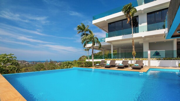 A modern hotel with a pool, palm trees, and a view of the ocean. - Baan Chom View Kamala (Kamala)