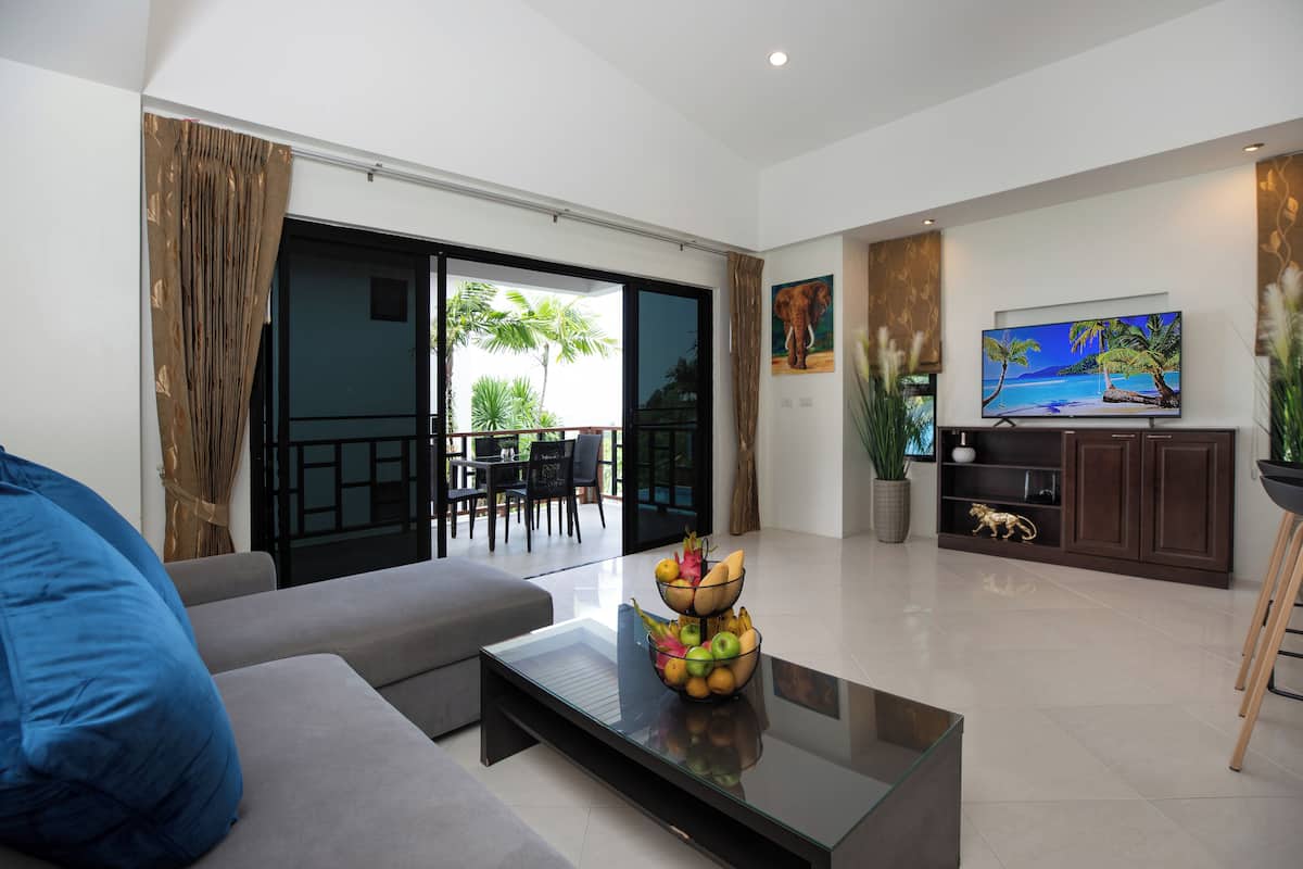A modern living room with a sofa, a coffee table with fruit, a TV, and a balcony with a view of palm trees.