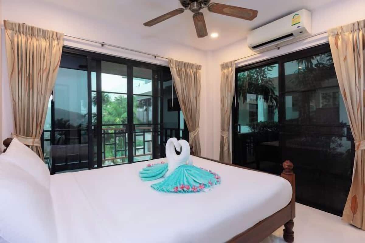 A hotel room with a bed, a ceiling fan, an air conditioning unit, and large windows with curtains.
