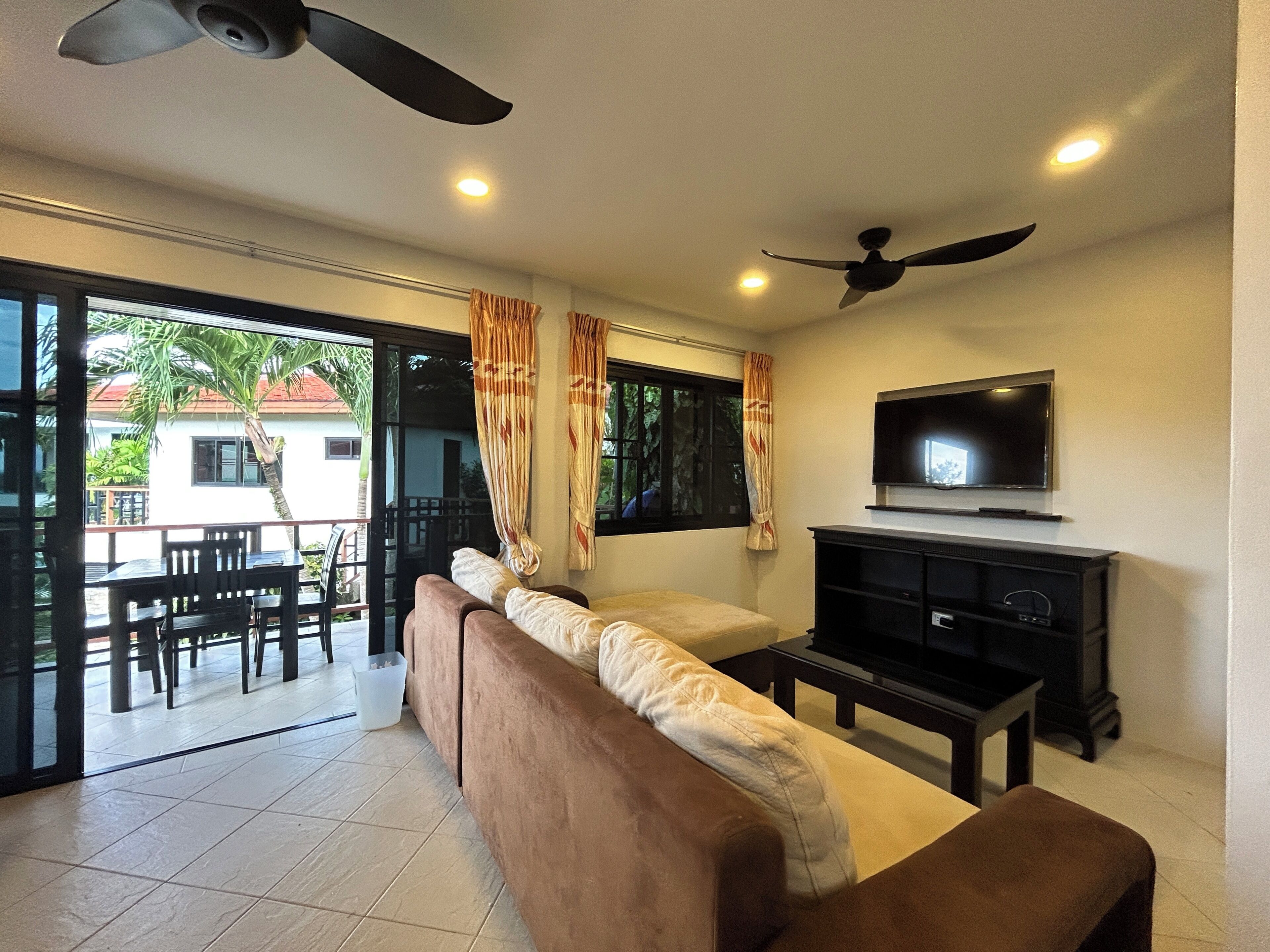 A living room with a sofa, TV, ceiling fan, and a view through a glass door to an outdoor area with a table and chairs.