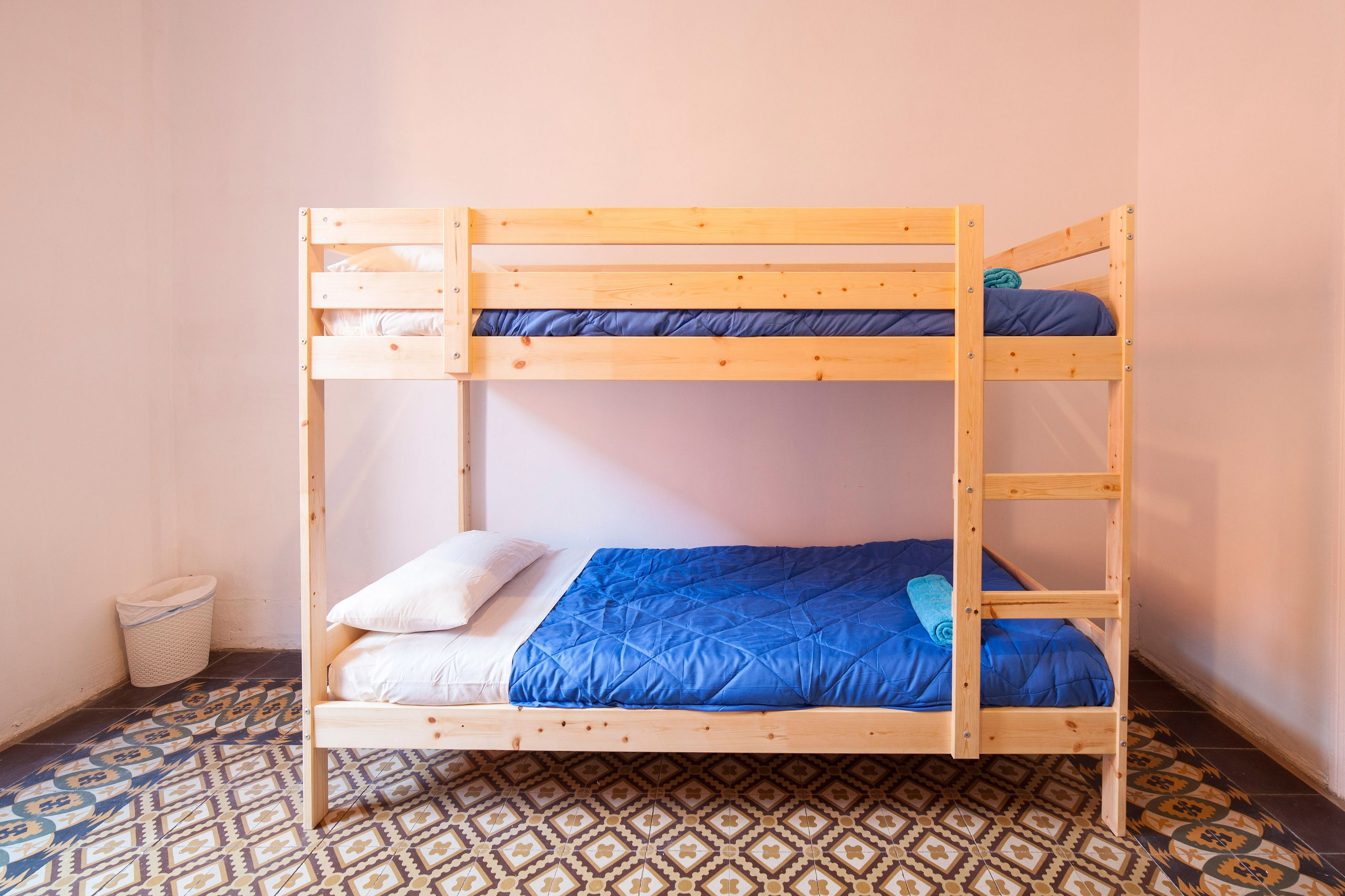 Una cama litera con colcha azul y almohadas blancas, un cubo de basura pequeño y baldosas de suelo estampadas.