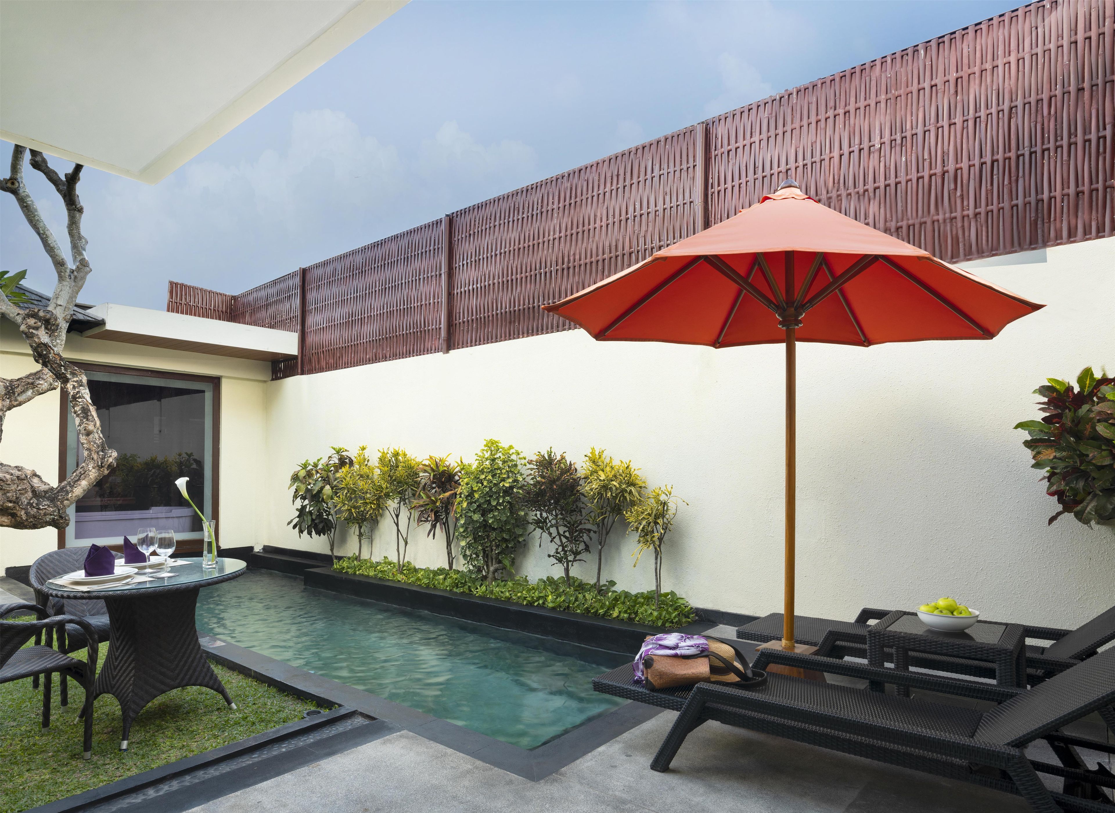 Un área junto a la piscina con sillones de descanso, una mesa de vidrio y una sombrilla roja.