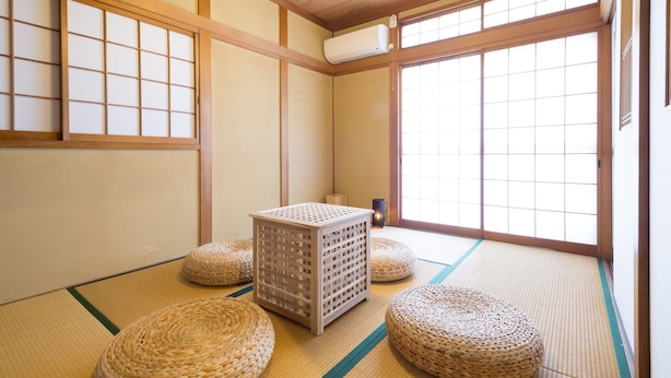A traditional Japanese room with tatami flooring, a tatami mat in the center, and a wooden air conditioning unit on the wall. - Sano san chi (Izumisano)