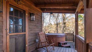 A wooden porch with a hot tub, a rocking chair, and a door with a glass panel. - Sunrise Cabin- Hot Tub, WiFi, Firepit, Grill (Campton)