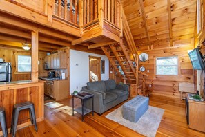 A wooden cabin interior with a kitchen, living area, and a staircase. - Sunrise Cabin | Hot Tub, WiFi, Firepit + Grill (Campton)