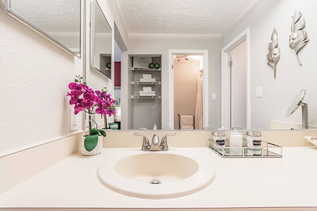Un baño con lavamanos blanco, espejo, florero verde con flores y toallero.