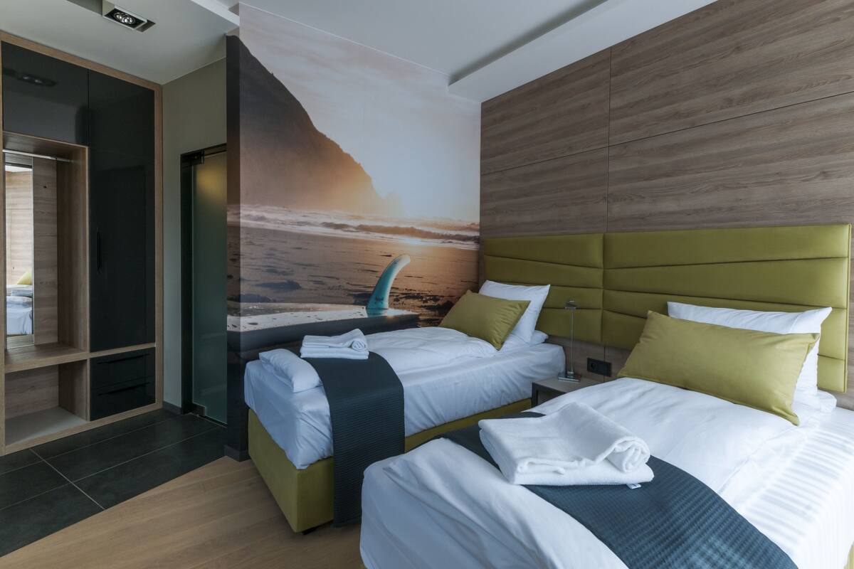 Une chambre d’hôtel avec deux lits, une tête de lit en bois, une fresque représentant une scène de plage et une armoire en bois.