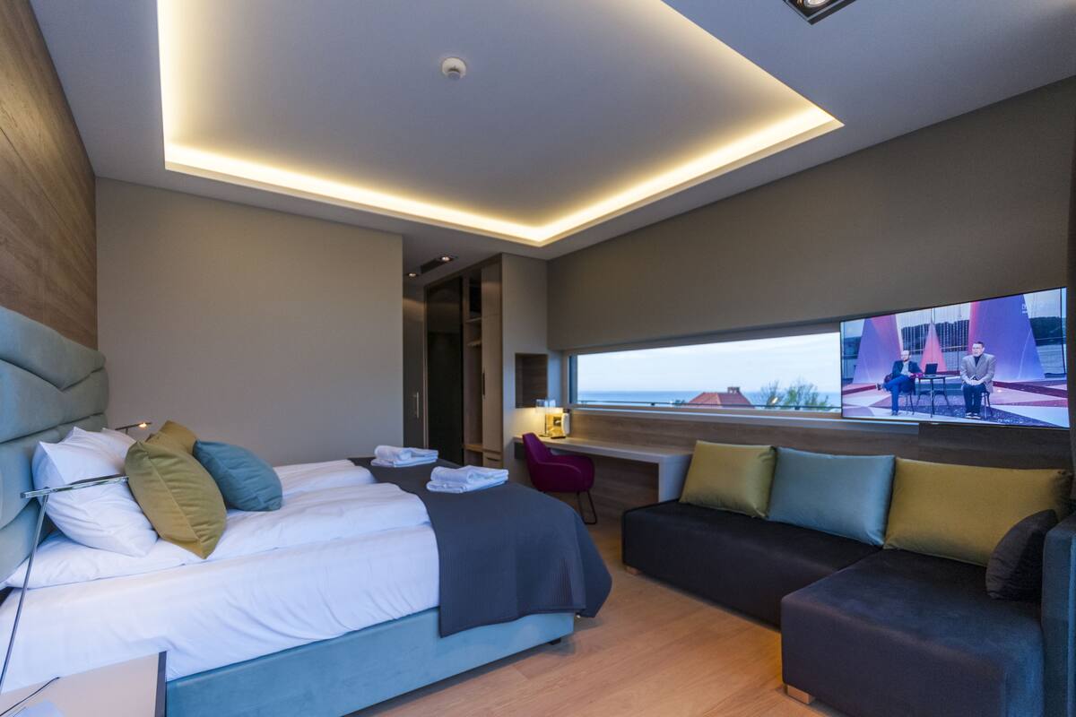 Une chambre d’hôtel moderne équipée d’un lit, d’un bureau, d’un canapé et d’un téléviseur diffusant des images d’un paysage pittoresque.
