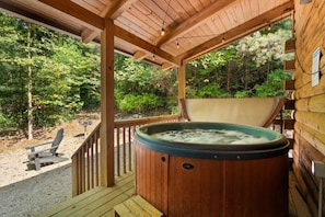 Une terrasse en bois avec un jacuzzi, une chaise et un chalet en bois, le tout situé dans une zone boisée.
