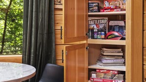 A cozy room with a wooden cabinet containing board games like Battleship and UNO. There's a table with chairs, a window with curtains, and a view of greenery outside.