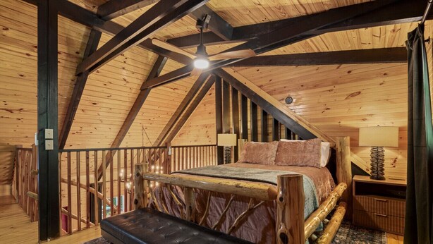 Un dormitorio acogedor en un  ático con techo de madera, una cama, un banco y un ventilador de techo.