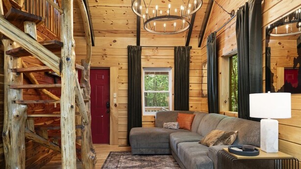 A log cabin interior with a wooden staircase, a gray sectional sofa, a patterned rug, and a chandelier.