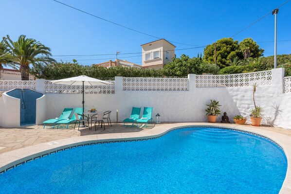 A swimming pool with a tiled deck, surrounded by a white wall with decorative patterns, and a small patio area with a table and chairs.