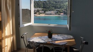 Une pièce dotée d’une grande fenêtre donnant sur une plage et des collines. On y trouve une table avec des chaises et une plante en pot.