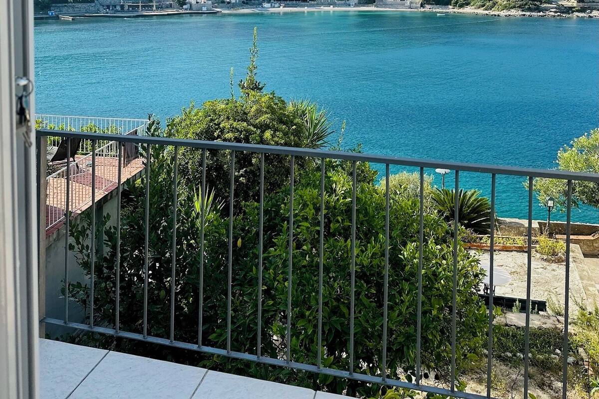 Une vue sur la côte, avec un balcon donnant sur une mer turquoise, une colline verdoyante et une rangée de bâtiments blancs.