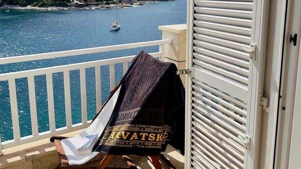 Un balcon avec vue sur la mer et un voilier, une chaise en bois et un barbecue avec une serviette.