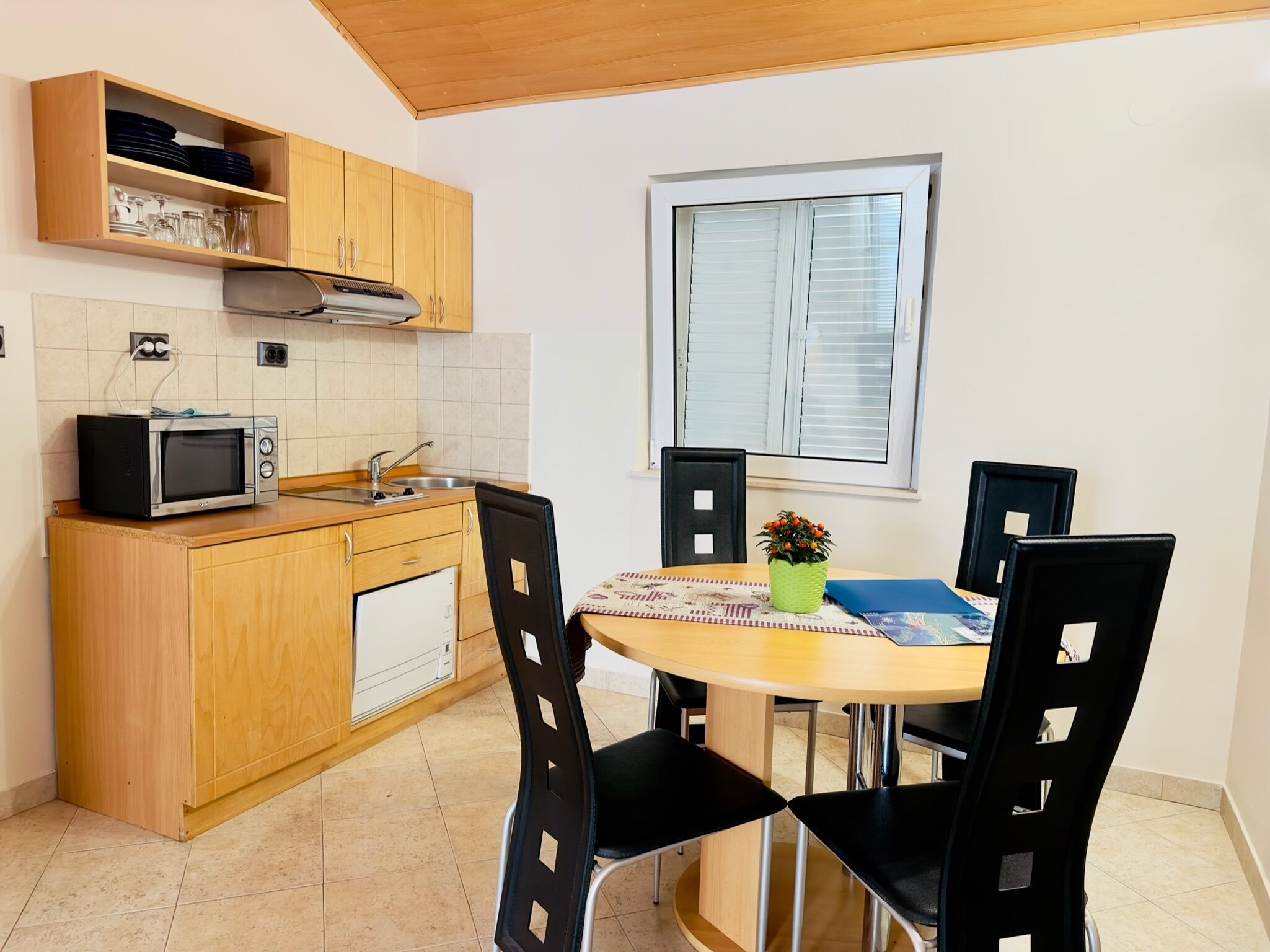 Cocina compacta con microondas, pileta y armarios de madera. Hay una mesa de comedor con sillas y un ventanal arriba.