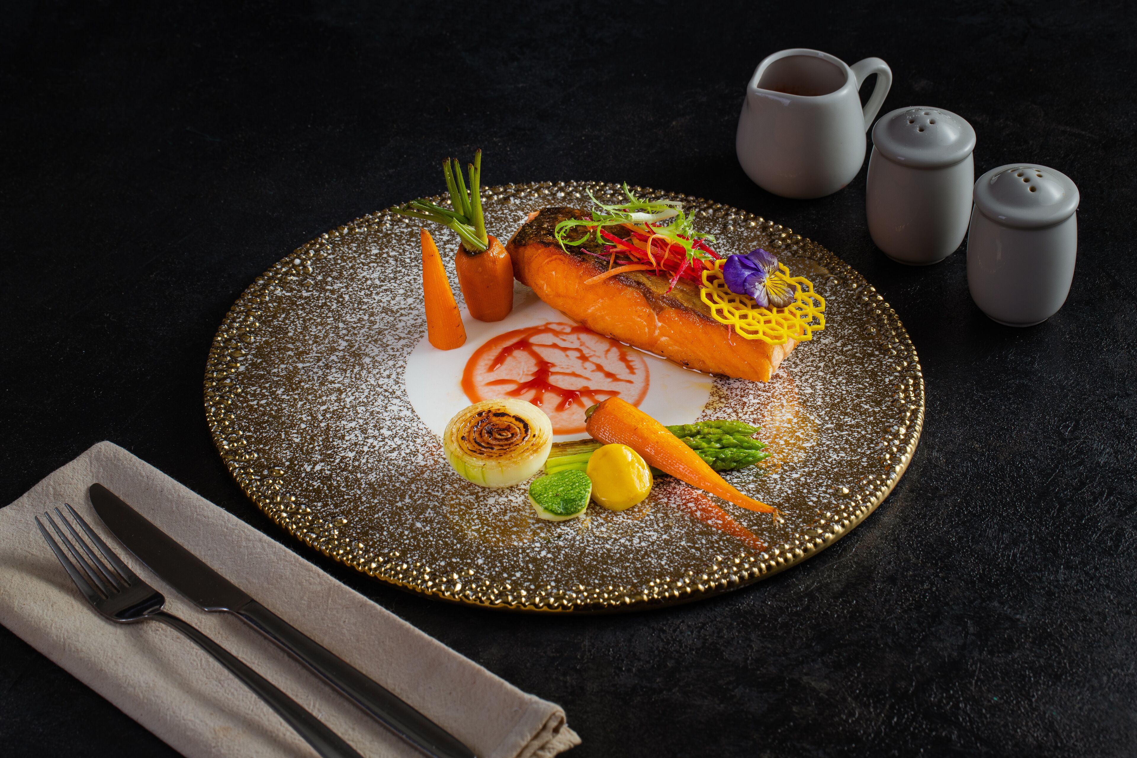 A plate of food with vegetables and a decorative garnish, accompanied by a napkin and cutlery on a dark surface.