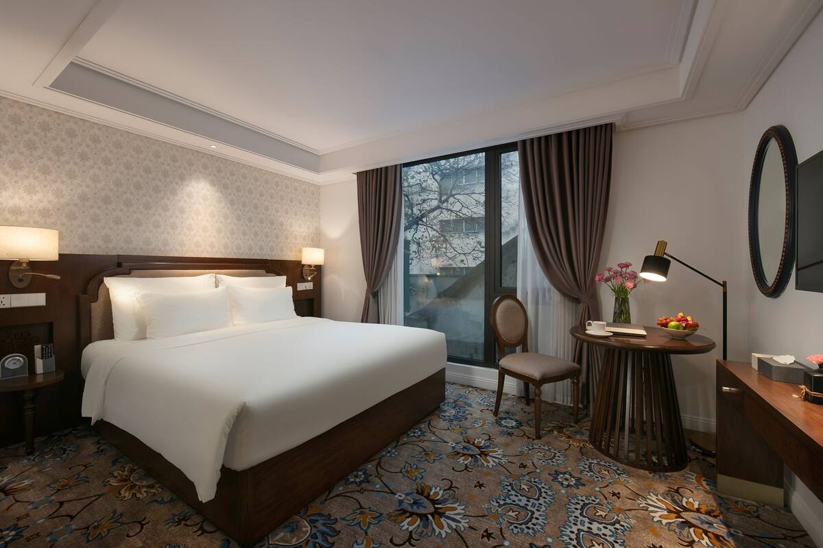 Habitación de hotel con una cama grande, un escritorio, una silla, una mesita con lámpara y un amplio ventanal con cortinas.