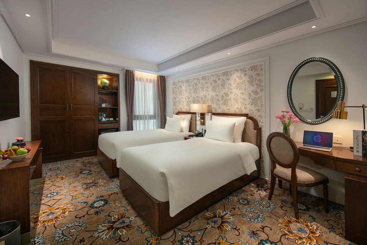 Habitación de hotel con dos camas, un escritorio, una silla, una computadora portátil, un espejo y una alfombra decorativa.