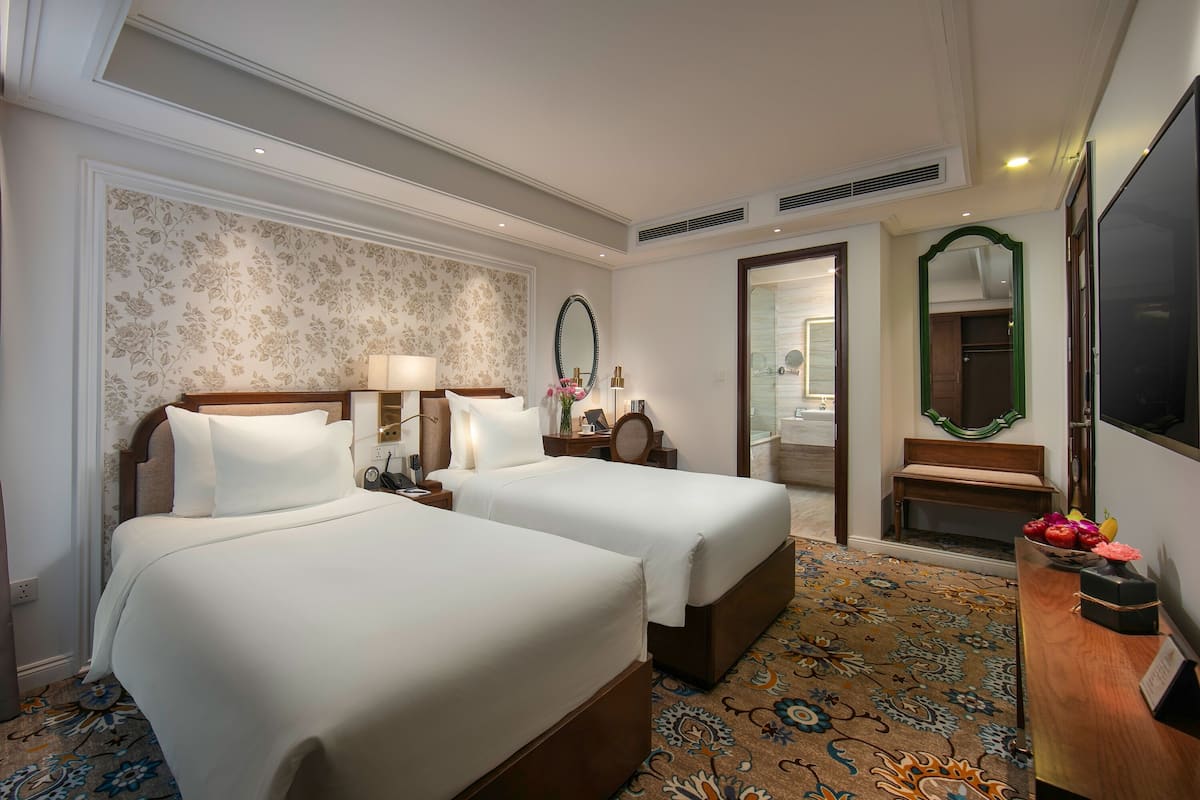 Habitación de hotel con dos camas, una zona de estar con un banco, un espejo, un televisor y un baño visible a través de una puerta abierta.