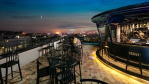 Un bar en la azotea con suelo de piedra, sillas de metal negro y una barra con vistas al horizonte de la ciudad al anochecer.
