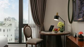 Quarto de hotel com uma janela grande, uma mesa redonda com um laptop, uma cadeira e um vaso com flores.