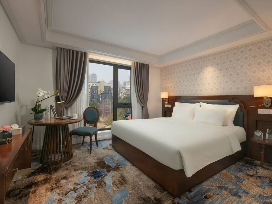 Habitación de hotel con una cama grande, un escritorio con silla, televisión y vista a la ciudad.