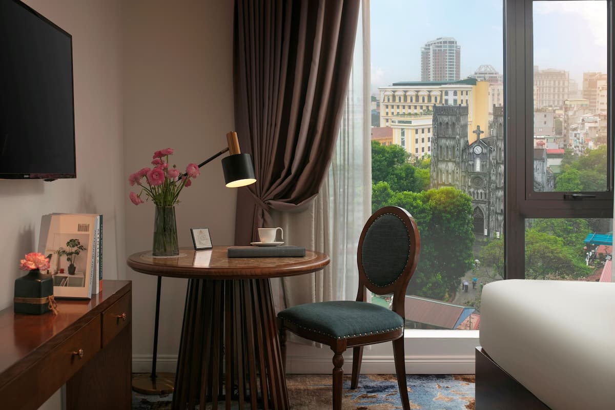 Una habitación de hotel con un ventanal grande, una mesa redonda de madera con una lámpara, una silla, una cama y un televisor en la pared.