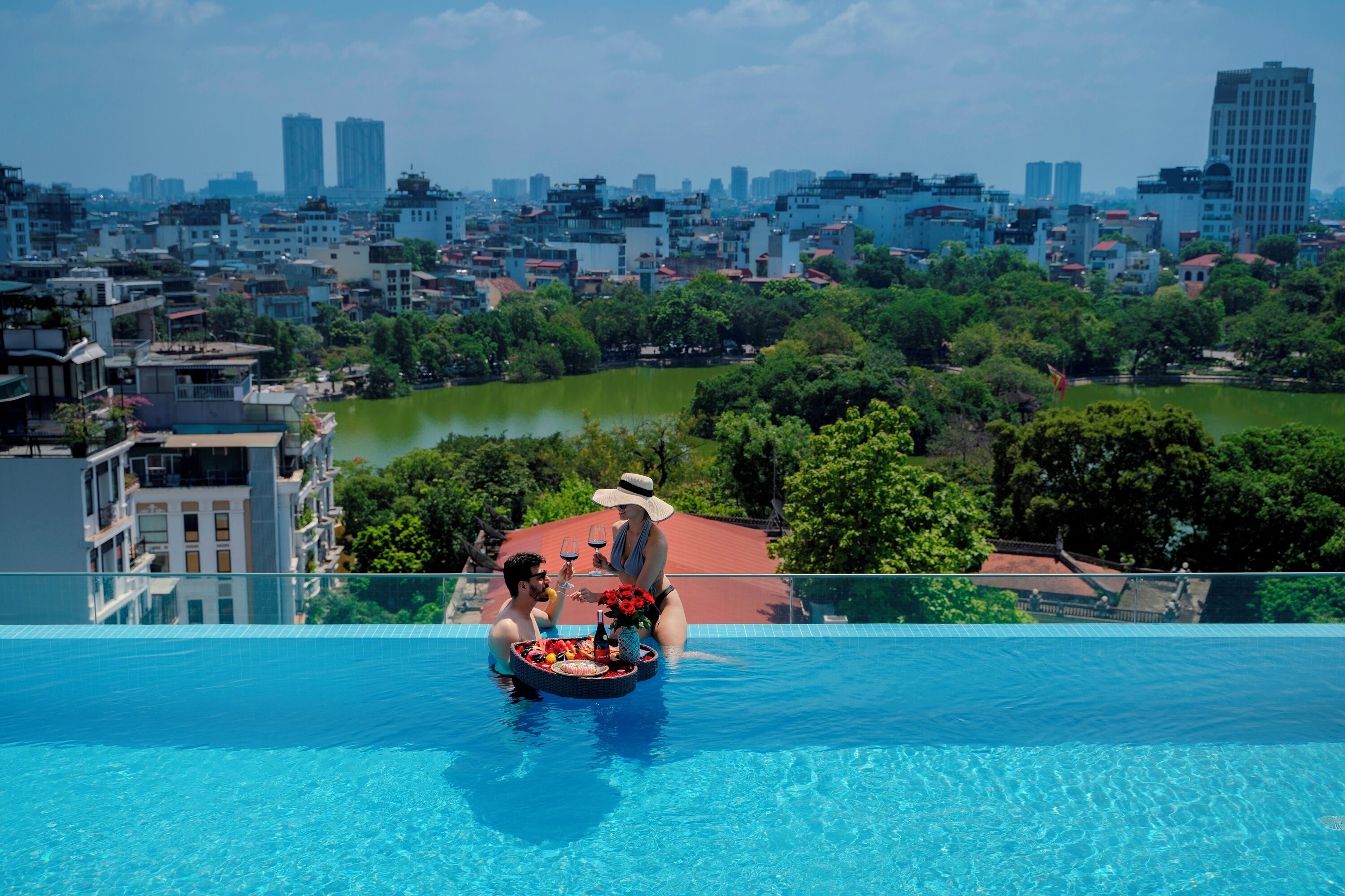 A rooftop pool with a cityscape view, a dining setup with a tray of food and drinks, and a person in a hat.