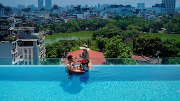 A rooftop pool with a cityscape view, a dining setup with a tray of food and drinks, and a person in a hat.