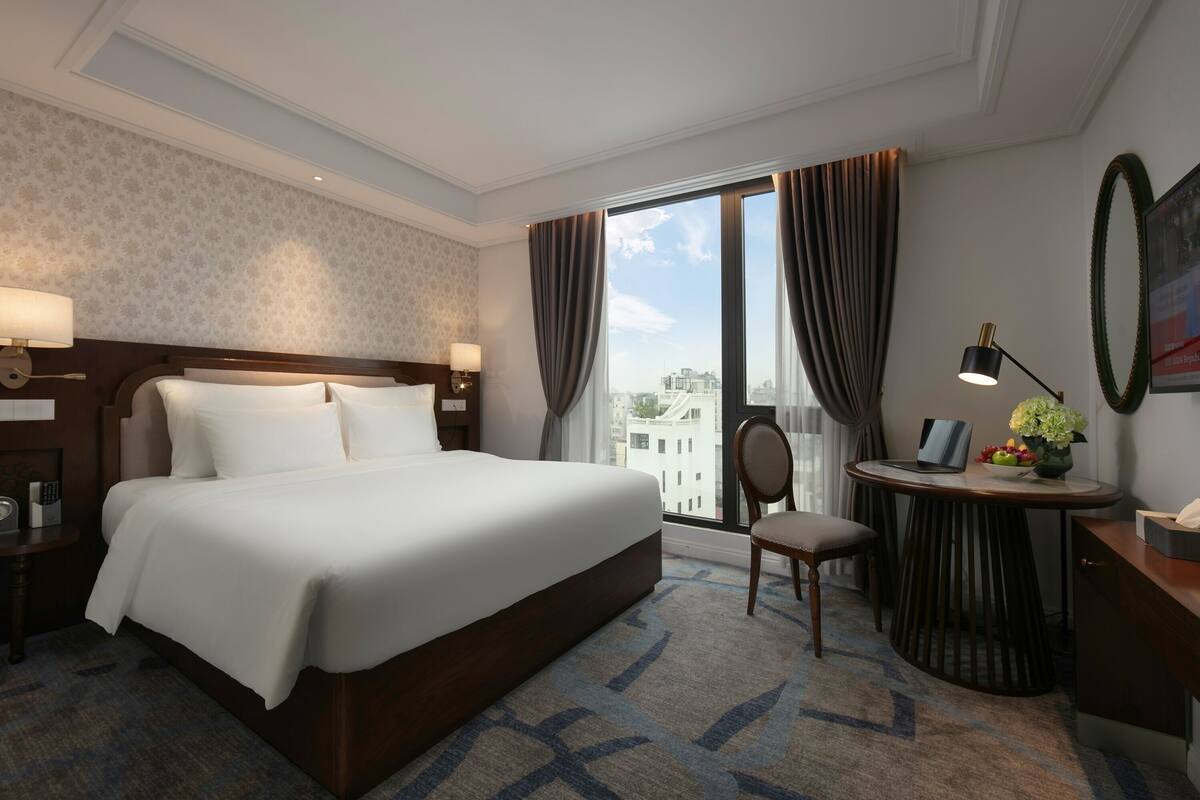 Habitación de hotel con una cama grande, un escritorio con lámpara, una silla y vistas de la ciudad a través de amplias ventanas.