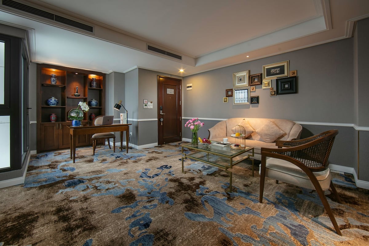 Una sala de estar con sofá, una mesa de centro y un tapete estampado. Se observa una estantería con objetos decorativos y una puerta con un letrero.